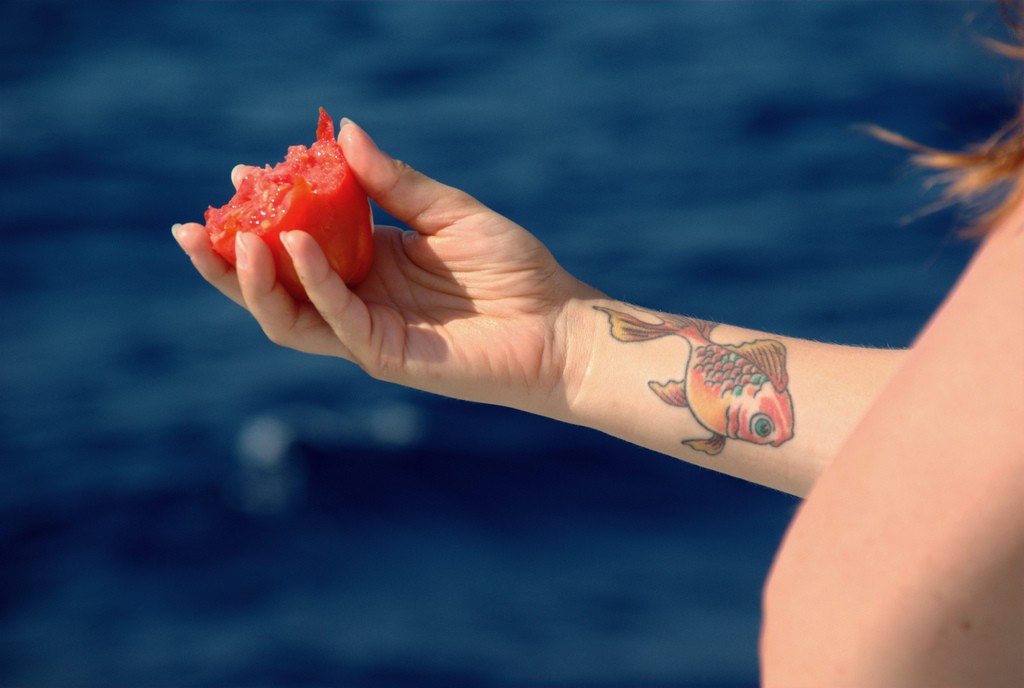A woman holds a half-eaten tomato. She has a goldfish tattoo on the inside of her wrist.