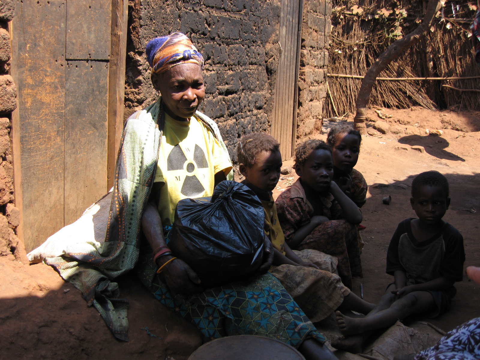 Older African woman sitting on the ground by a rough, brick building and a black plastic bag in her lap, surrounded by four young children.