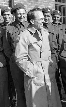 A black and white photo of Tommy Douglas, a middle aged man with glasses. Four military men are standing in the background.