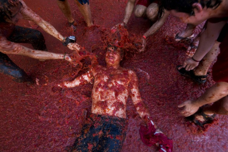 People standing in a tub of tomato pulp toss it onto a shirtless man lying down.
