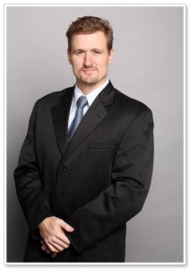 A broad man in a black suit with a blue tie and a small smile.