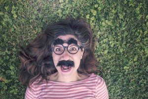 A woman lies on grass, wearing a fake moustache, big nose, glasses, and bushy eyebrows.