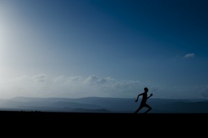 A runner is silhouetted against a landscape of mountains and blue sky with clouds.