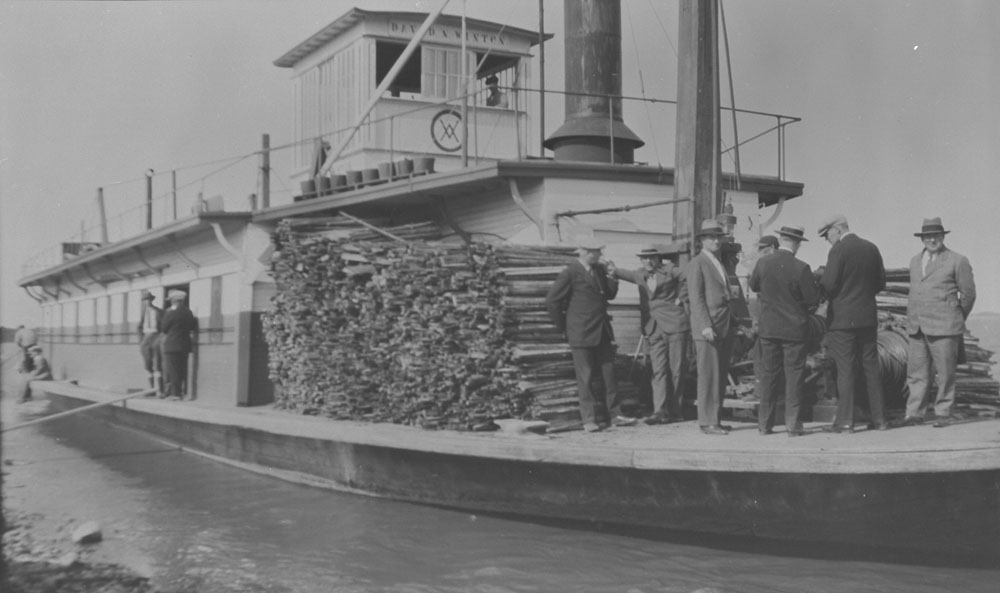 A steamer ship with a dozen men in suits and a large pile of lumber on the deck.