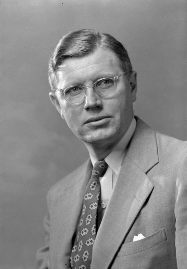 Close-up of a man in a suit with a patterned tie and clear-rimmed glasses.
