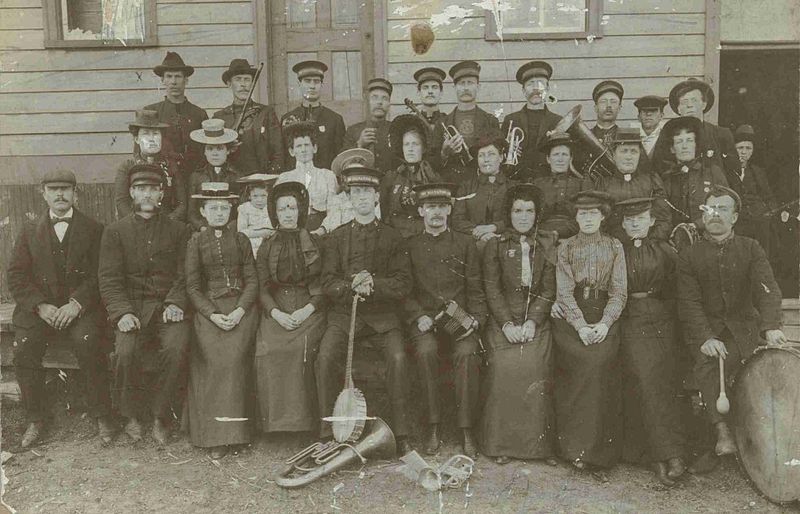 Three rows of men and women, some with musical instruments, pose in front of a wooden building.