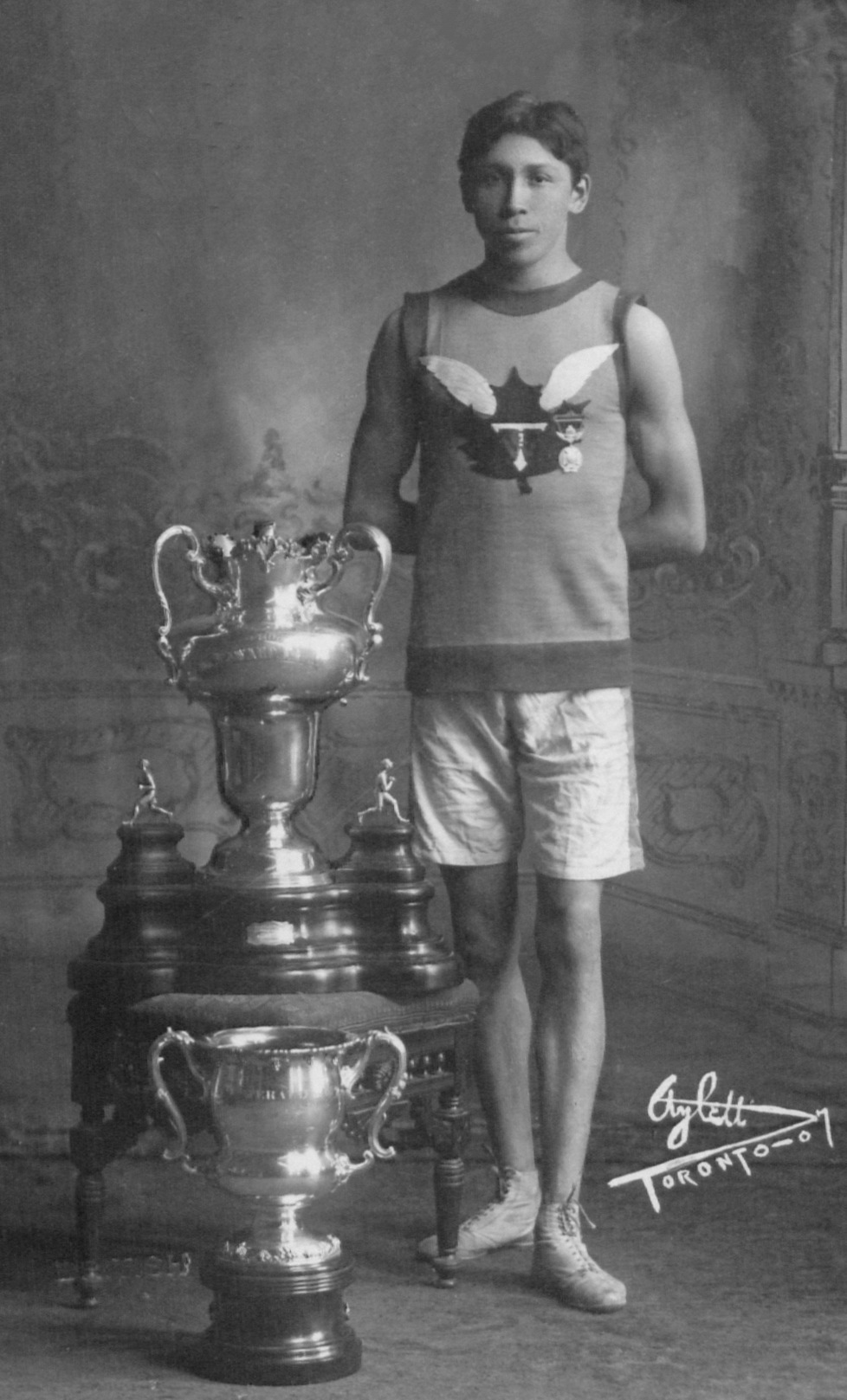 A young man poses beside two large trophies. He wears shorts, a tank top, and running shoes.