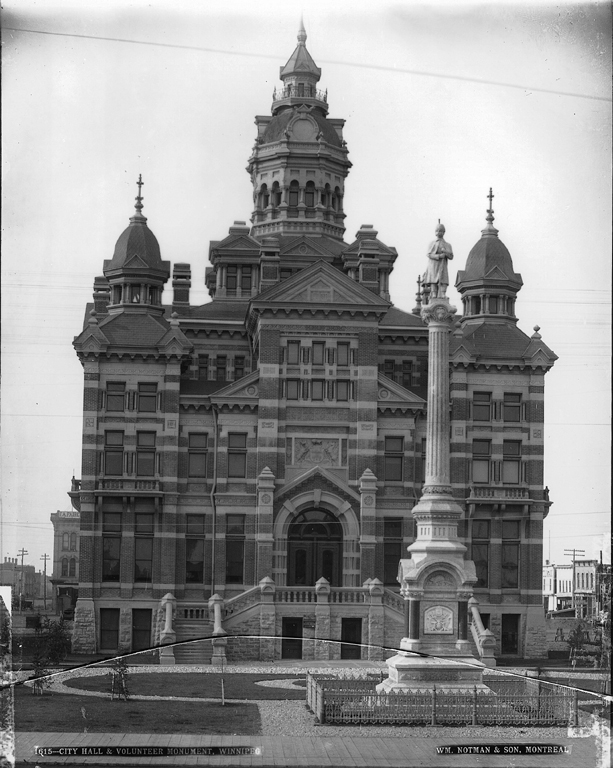 Tall, ornate city hall. In front is a tall, skinny monument with a statue of a man on top.