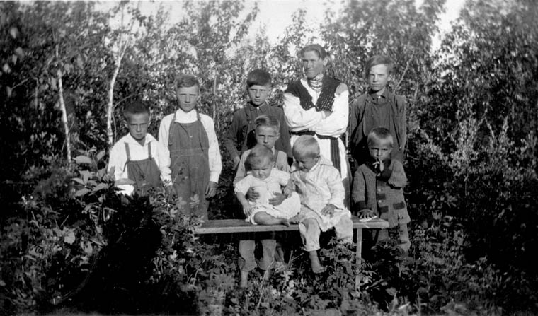A woman poses with eight children (mostly boys) in front of a stand of bushes.