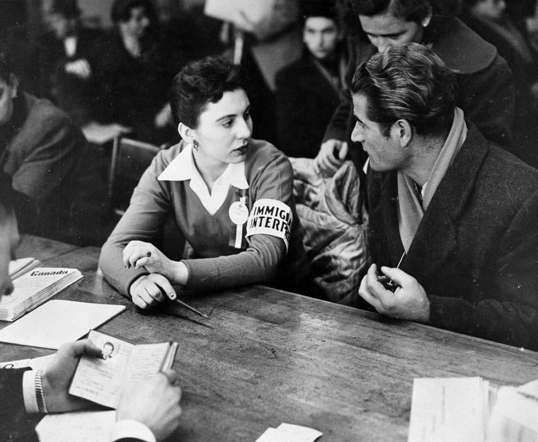 An immigration interpreter sits and talks with a man. A man across the table inspects a passport.