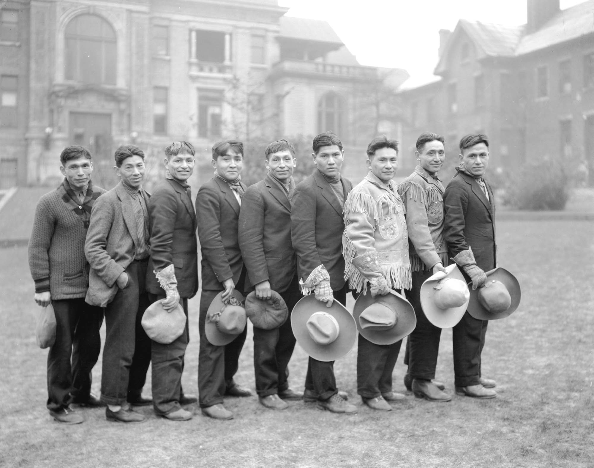 Nine men stand in a line, hats held in front of them, grinning.