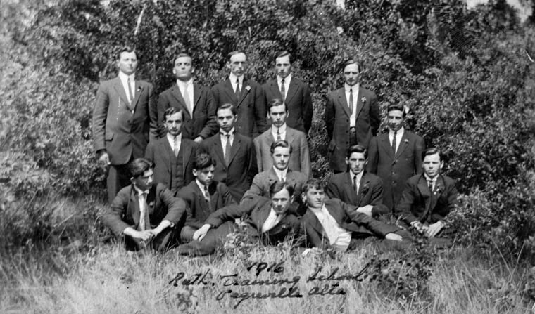 Sixteen young men in suits pose outdoors on grass in front of foliage.