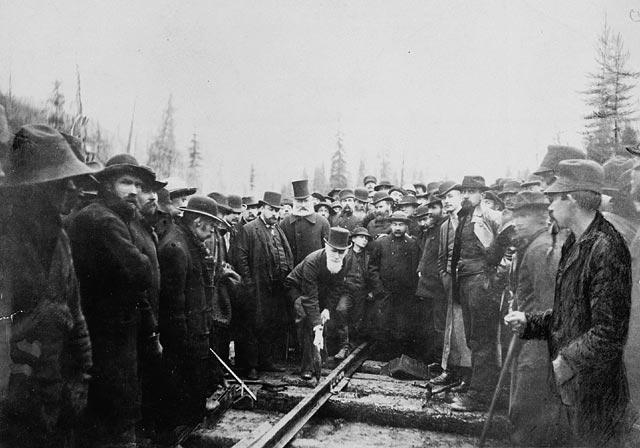 Railway workers stand around a man in a suit with a top hat who is hammering a spike into a railway.