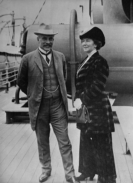 An older man in a suit and an older woman in a long dress stand on a ship deck.