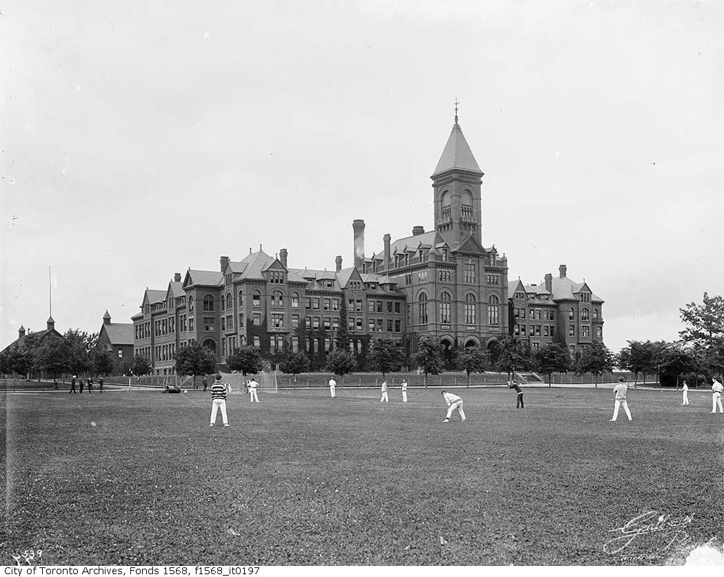 Young men play cricket outside of an ornate college building.