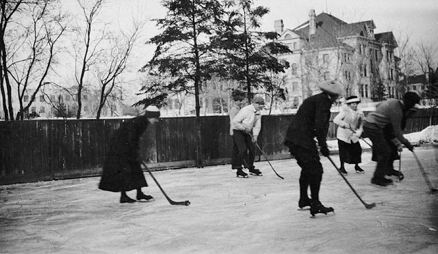 Men and women play ice hockey on a pond near a tall residential building.
