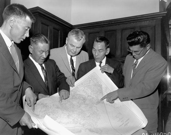Five men in suits hold a map and point at it.