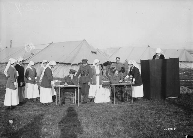 Several nurses gather around tables manned by officers; one is in a voting booth. Behind are tents.