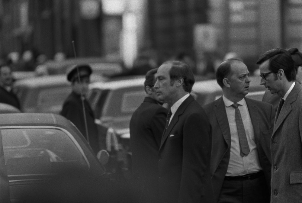 Two men in suits walk behind a sea of cars, looking solemn.
