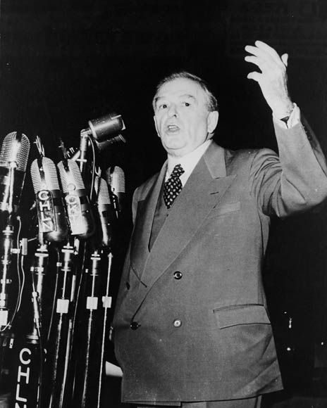 A middle-aged man in a suit gestures while speaking into several microphones.