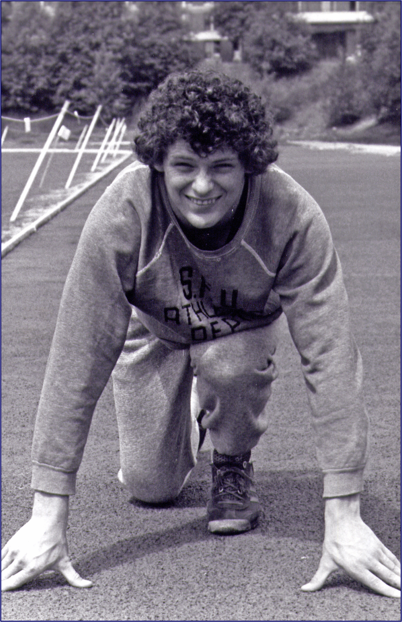 A smiling young man with curly hair wearing a tracksuit poised for a crouch start.