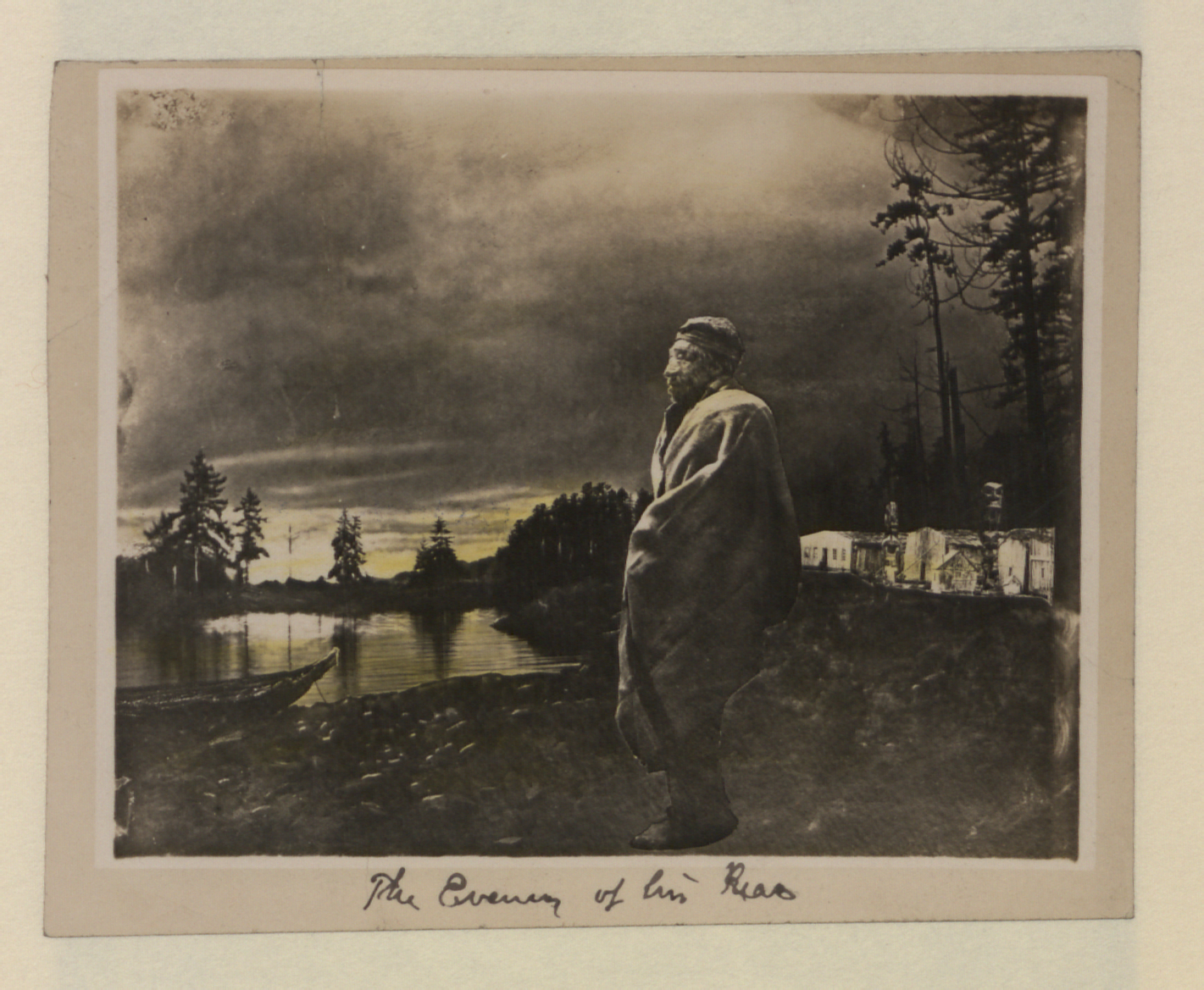 An Indigenous man stands wrapped in a blanket. A river is behind him, a canoe sitting on the bank.