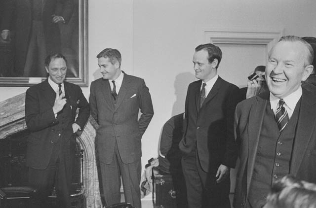 Four smiling men in suits stand in a room with white walls. An ornate portrait hangs behind them.