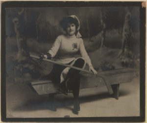 A woman sits on a wooden bench on an ice rink, holding a hockey stick across her knee.