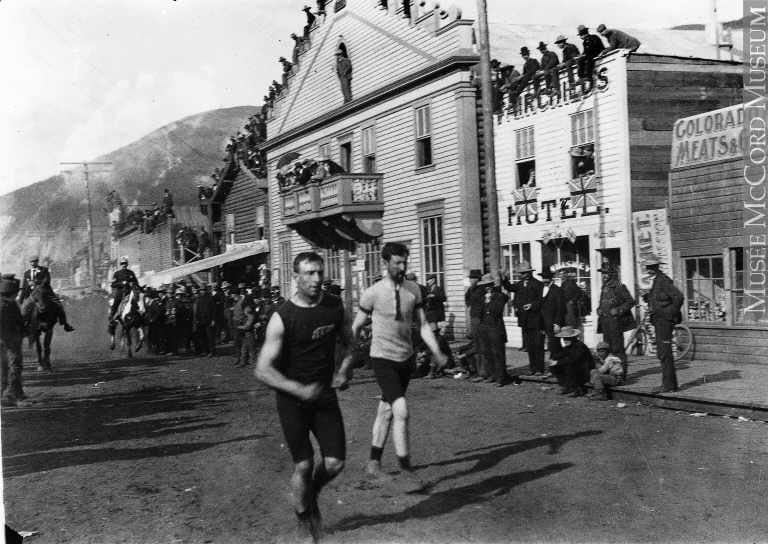 Two men run past a crowd gathered outside simple storefronts.