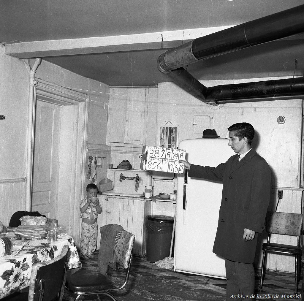 A man holds a numbered sign in a kitchen. A shy boy in the corner drinks from a mug.