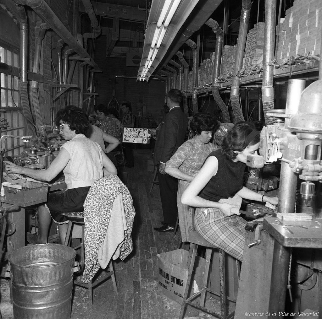 Women work at stations in a factory. One woman's faux leopard coat is over the back of her chair.