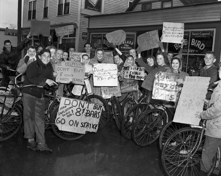 A crowd of children with their bikes, holding signs protesting 8-cent candy bars.