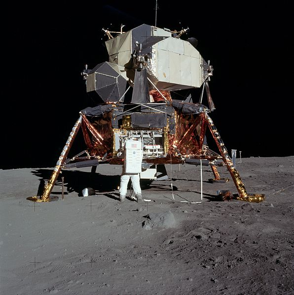 A spacecraft with legs and a metal capsule. An astronaut stands in front of it.