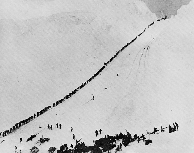 A long line of people ascend a snowy mountain pass.