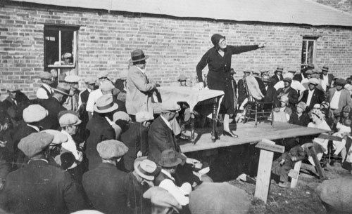 A woman stands on a table, addressing a crowd outside a low brick building.