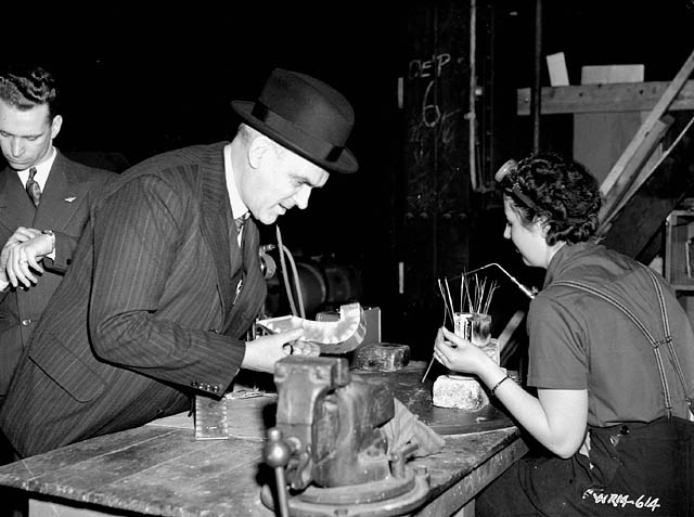 A man in a suit and a fedora inspects a piece of metal with a female factory worker looking on.