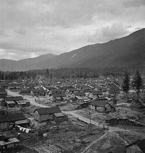 Collection of low buildings at the foot of a mountain, beside a forest.