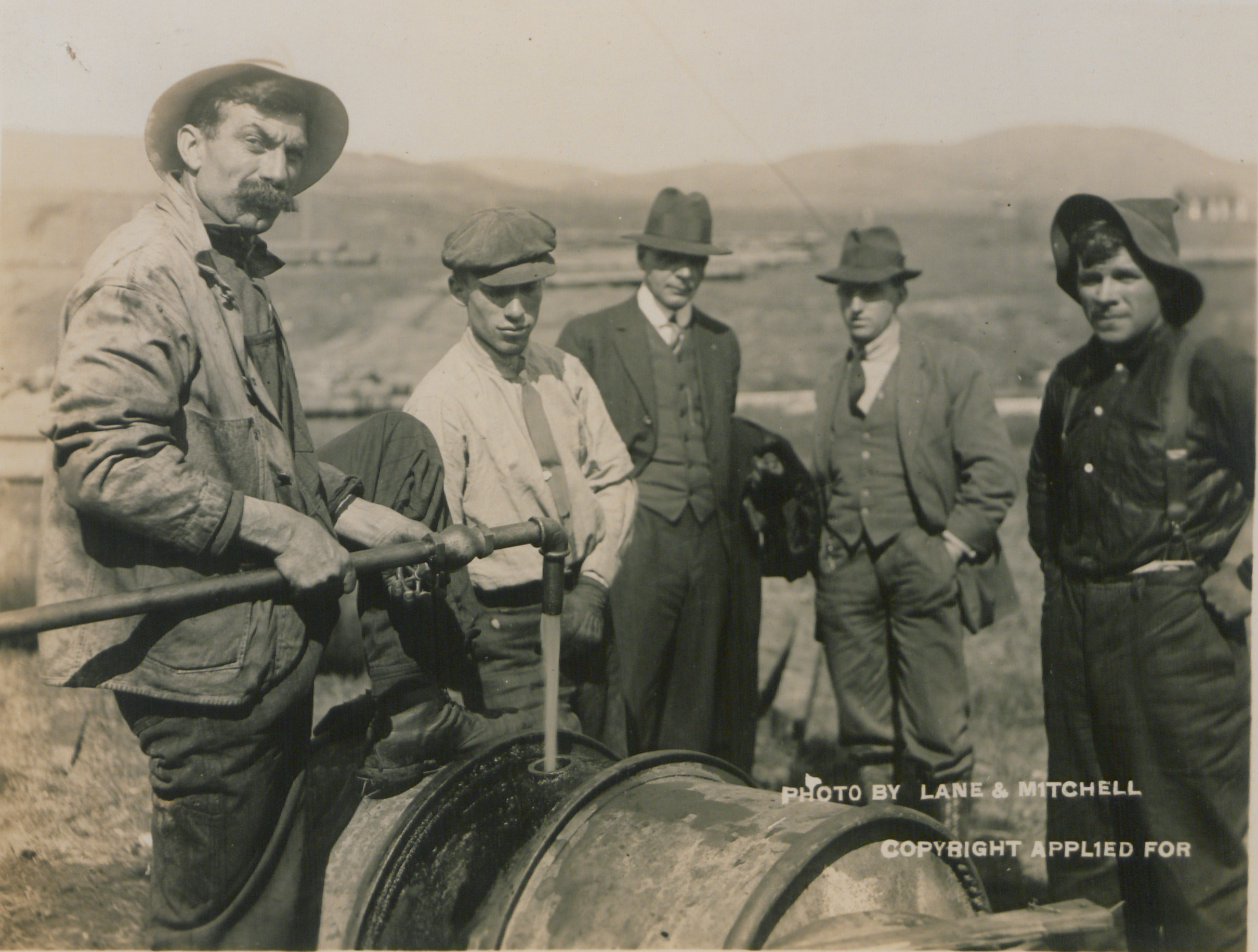 A man feeds oil into a drum using a pipe. Four other men (two working, two in suits) look on.