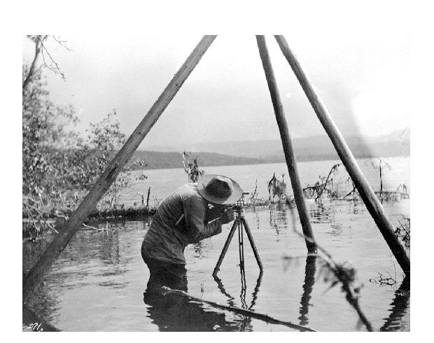A man stands in the shallows of a lake, bent over a tripod.
