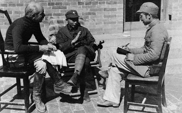 Three men sit and talk on mismatched chairs on a stone patio.