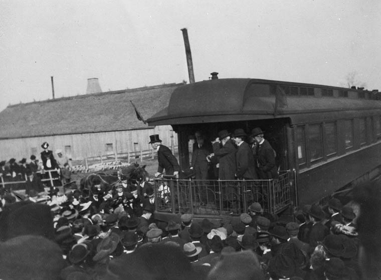 A crowd gathered around a railway car. Five men stand on the platform of the observation car. 