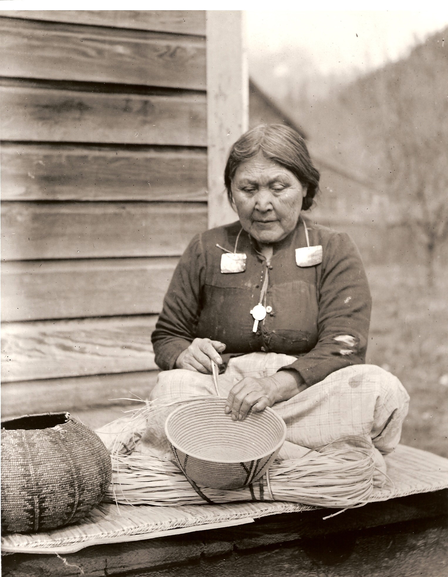 An older woman sitting on a porch weaves a basket.