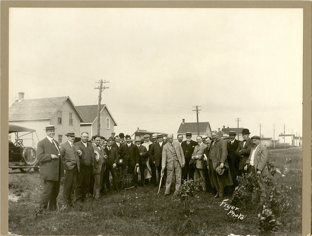 Two dozen men pose in suits, with one in the centre holding a shovel in the ground.