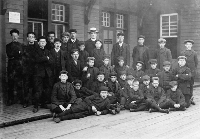 Thirty boys pose outside of a wooden building.