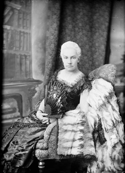 A woman wearing a Victorian gown sits in an opulent armchair, fanning herself.