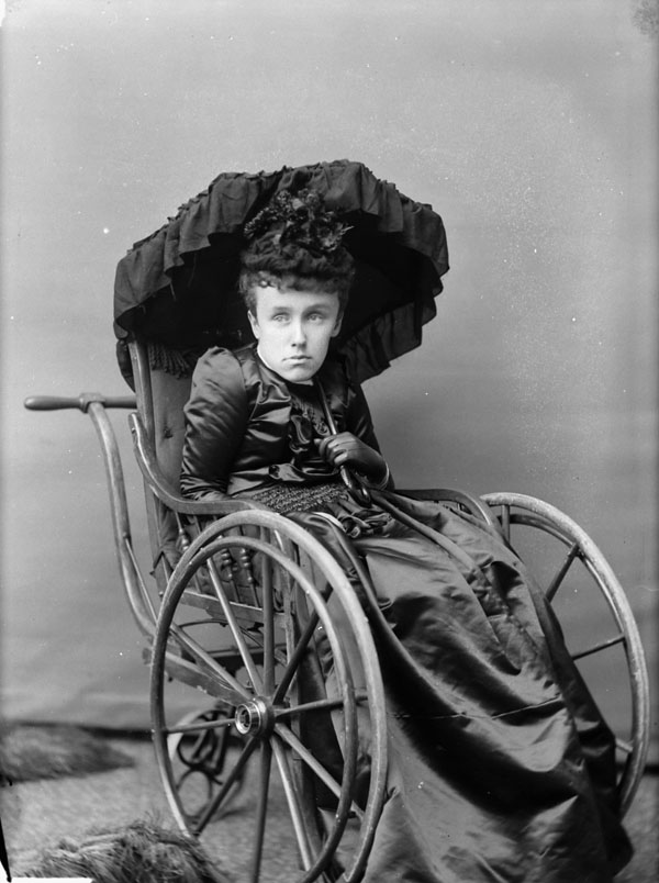 Kobieta w ciemnej sukience trzyma czarny parasol i siedzi na wózku inwalidzkim.