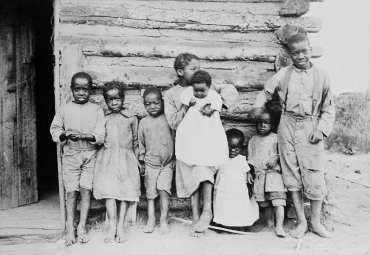Eight children of various ages stand outside a log cabin.