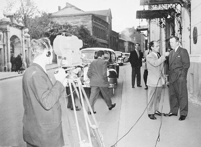 A TV camera is pointed at a man interviewing another man. A couple of men look on.