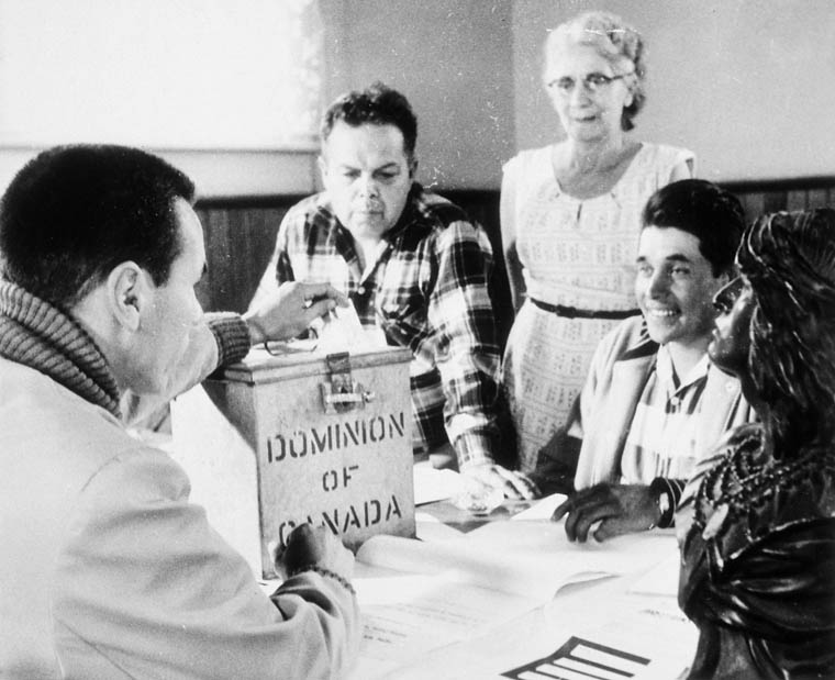 Three men and a woman are gathered around a Dominion of Canada ballot box.