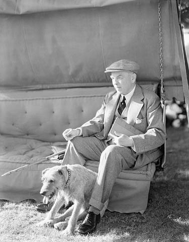 A middle-aged man wearing a suit sits on an outdoor couch with a terrier at his feet.